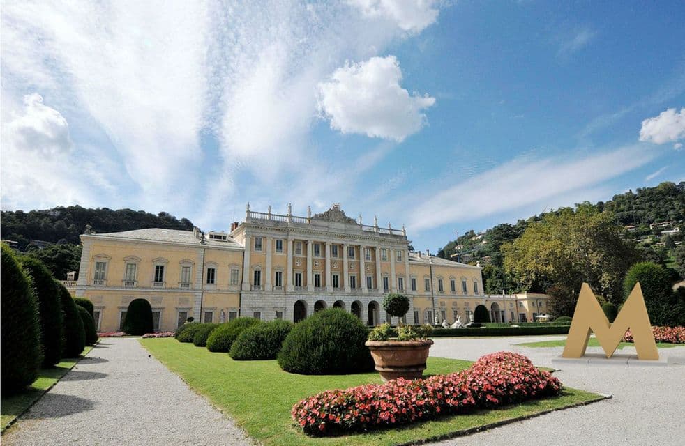 Una grande villa classica gialla si erge dietro giardini curatissimi, con un sentiero di ghiaia e una grande scultura a 'M' dorata.