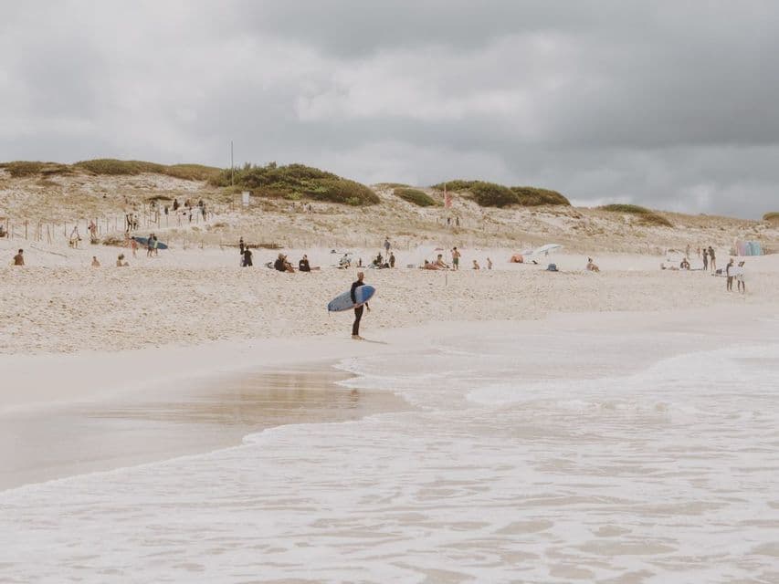 Un surfista in muta tiene una tavola da surf blu a riva su una spiaggia sabbiosa sotto un cielo nuvoloso.