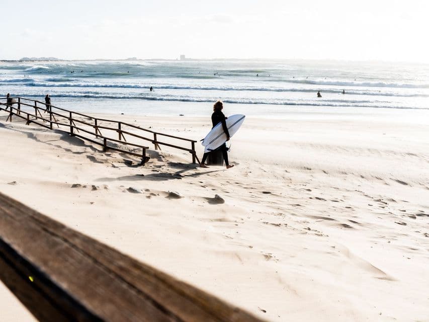 Un surfista in muta trasporta una tavola da surf e cammina a piedi nudi su una spiaggia sabbiosa verso l'oceano.