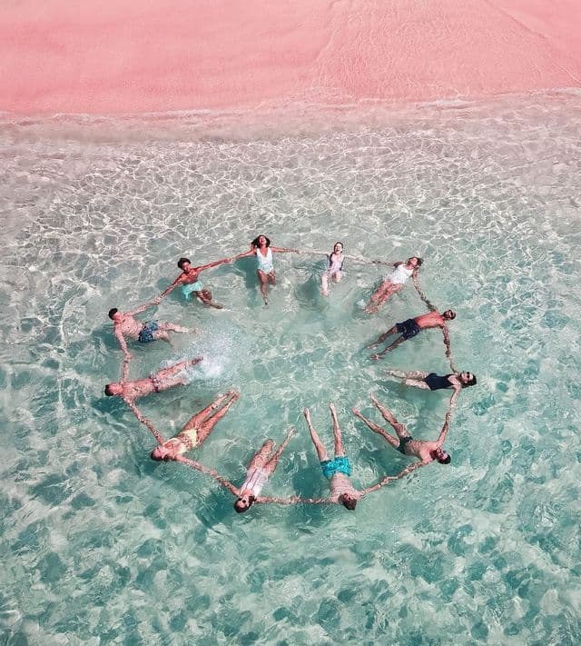 Vue aérienne d'un voyage de groupe WeRoad flottant en cercle, se tenant les mains dans l'eau claire près d'une plage de sable rose.
