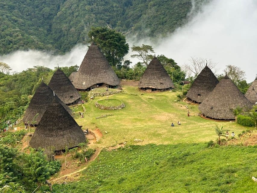 Une vue plongeante d'un village traditionnel avec des huttes coniques aux toits de chaume sur un flanc de montagne herbeux, avec des collines boisées et du brouillard en arrière-plan.