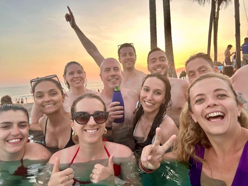 Un voyage de groupe WeRoad prenant un selfie dans une piscine au coucher du soleil, avec une plage et des palmiers en arrière-plan.