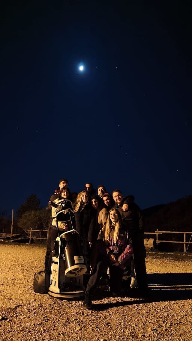 Un gruppo WeRoad in viaggio posa con un grande telescopio sotto un cielo notturno sereno e una luna brillante.