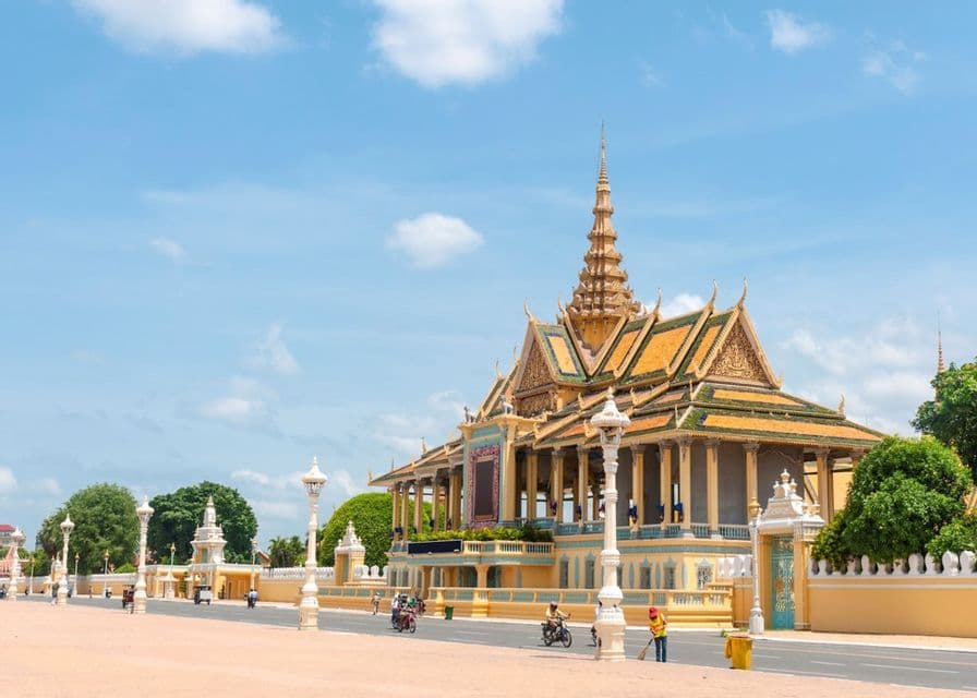 An ornate yellow palace with a multi-tiered golden roof stands beside a paved square under a bright blue sky.