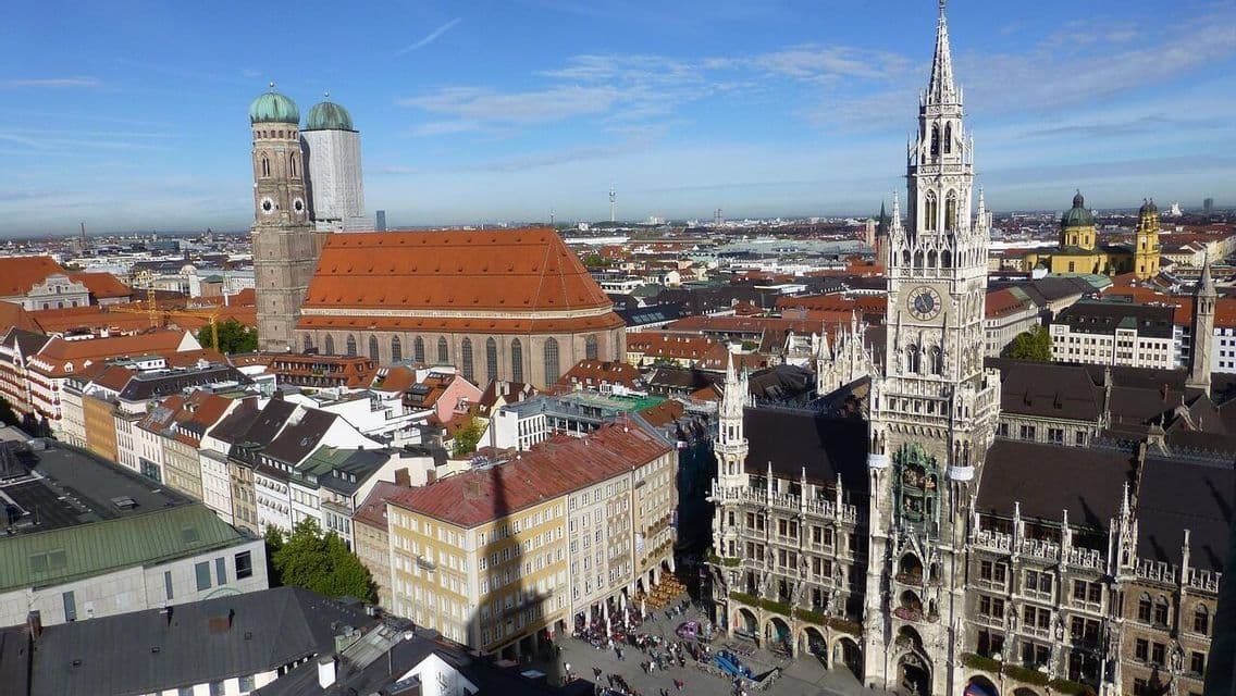 Eine Luftaufnahme einer europäischen Stadtlandschaft mit einem großen gotischen Uhrenturm und einer Kathedrale mit zwei Kuppeln unter blauem Himmel.
