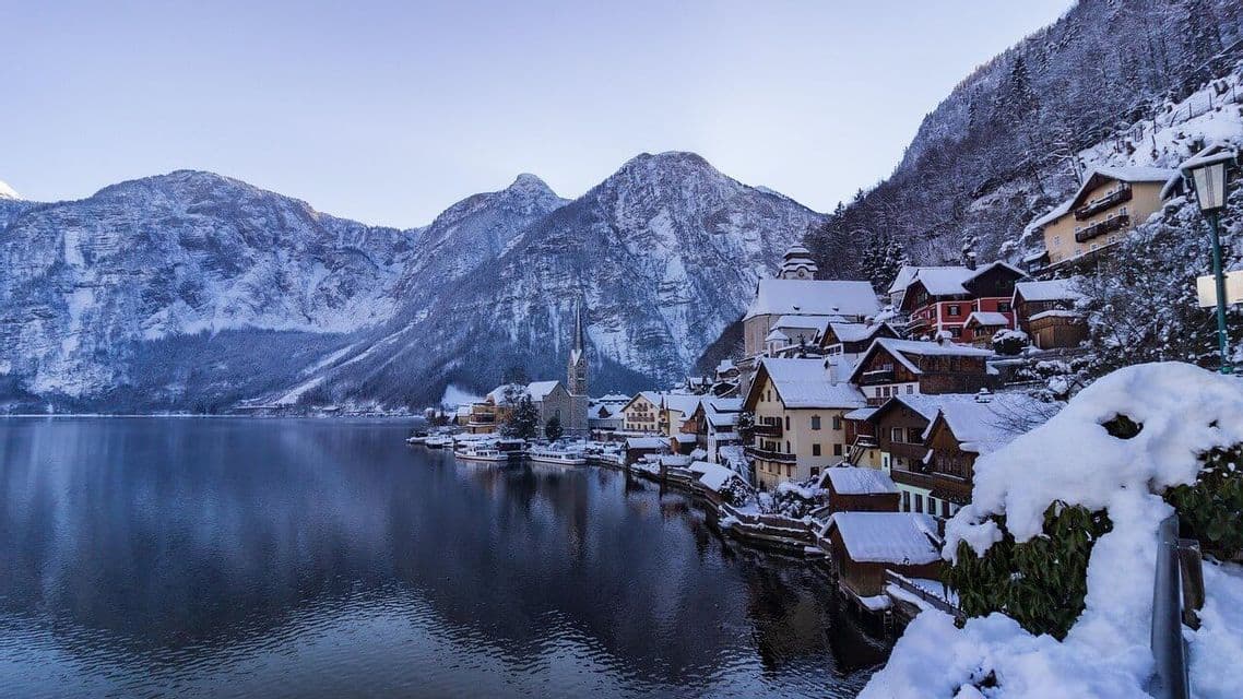 Ein schneebedecktes Dorf mit einer Kirche, eingebettet an einem ruhigen Seeufer am Fuße großer Berge unter einem klaren Himmel.