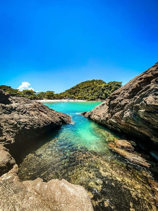 A rocky inlet with clear turquoise water opens to a sandy beach with people, backed by a lush green hill under a clear blue sky.
