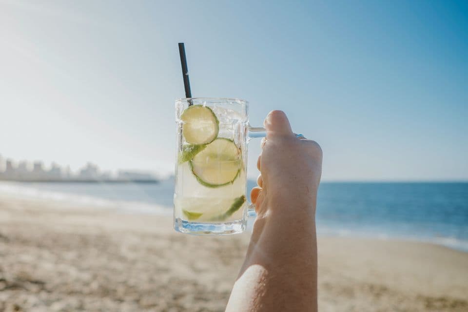 A hand holds up a glass mug filled with a lime cocktail and a black straw, with a sunny beach and the ocean in the background.