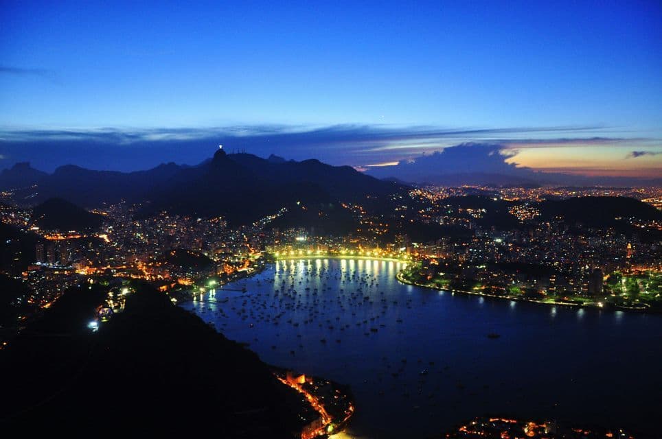 An aerial view of a city surrounding a bay filled with boats at dusk, with its lights on and a statue on a distant mountain.