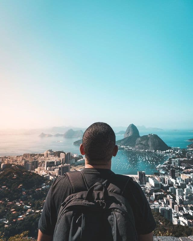 A person wearing a backpack looks out over a panoramic view of a coastal city and a bay with mountains.