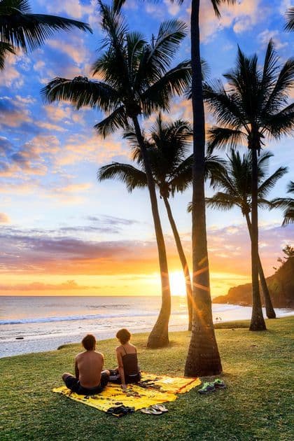 Deux personnes sont assises sur une couverture dans l'herbe, contemplant le coucher de soleil sur une plage bordée de grands palmiers.