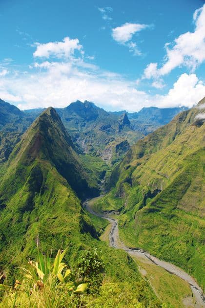 Vue panoramique d'une rivière sinuant à travers une vallée profonde entre des montagnes luxuriantes et vertes sous un ciel partiellement nuageux.