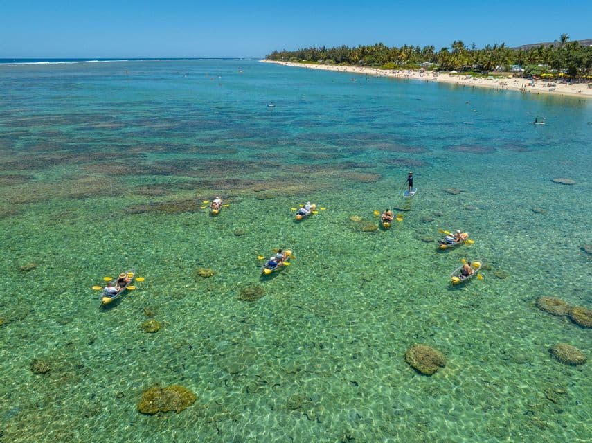 Vue aérienne d'un voyage de groupe WeRoad en kayak dans des bateaux transparents au-dessus d'un récif corallien clair près d'une plage de sable.