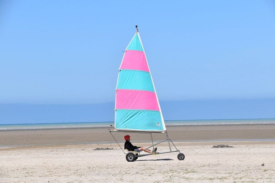 Une personne avec un casque rouge est assise dans un char à voile avec une voile rose et turquoise sur une large plage de sable.