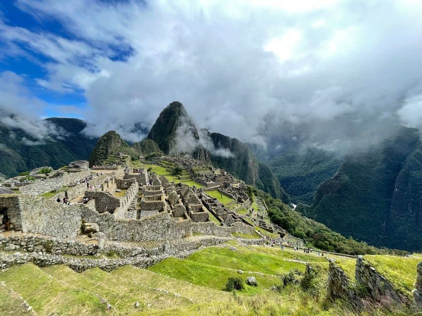 Ein weitläufiger Blick auf antike Steinruinen und grüne Terrassen an einem Berghang, mit wolkenverhangenen Gipfeln im Hintergrund.