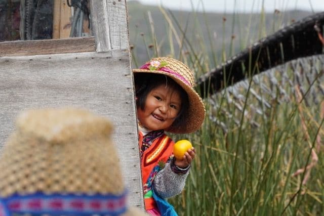 Una giovane ragazza sorridente con un cappello di paglia e vestiti colorati tiene un frutto giallo, spuntando da dietro una struttura di legno.