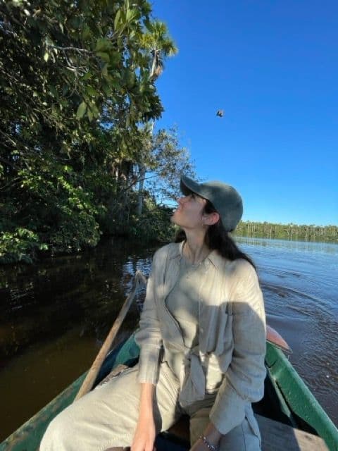 Una donna con un cappello siede in una piccola barca su un fiume calmo, guardando un uccello che vola nel cielo azzurro chiaro.