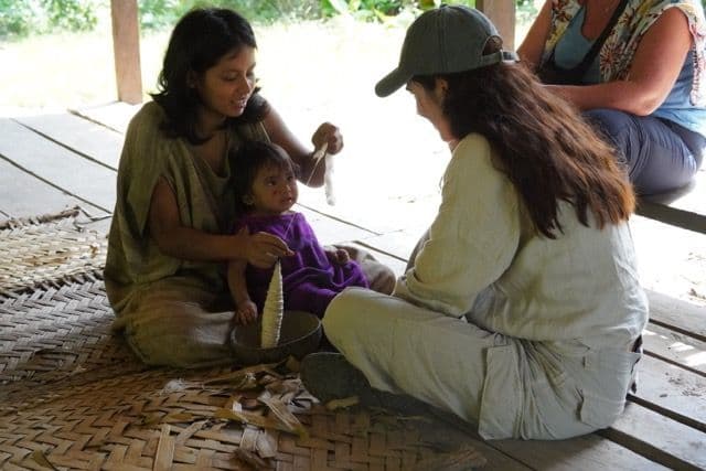 Una donna del posto con un bambino in grembo mostra a un viaggiatore di un viaggio di gruppo WeRoad come filare la fibra naturale su un tappeto intrecciato.