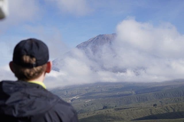 Una persona con un cappellino nero, vista di spalle, osserva un'imponente cima montuosa che emerge dalle nuvole.