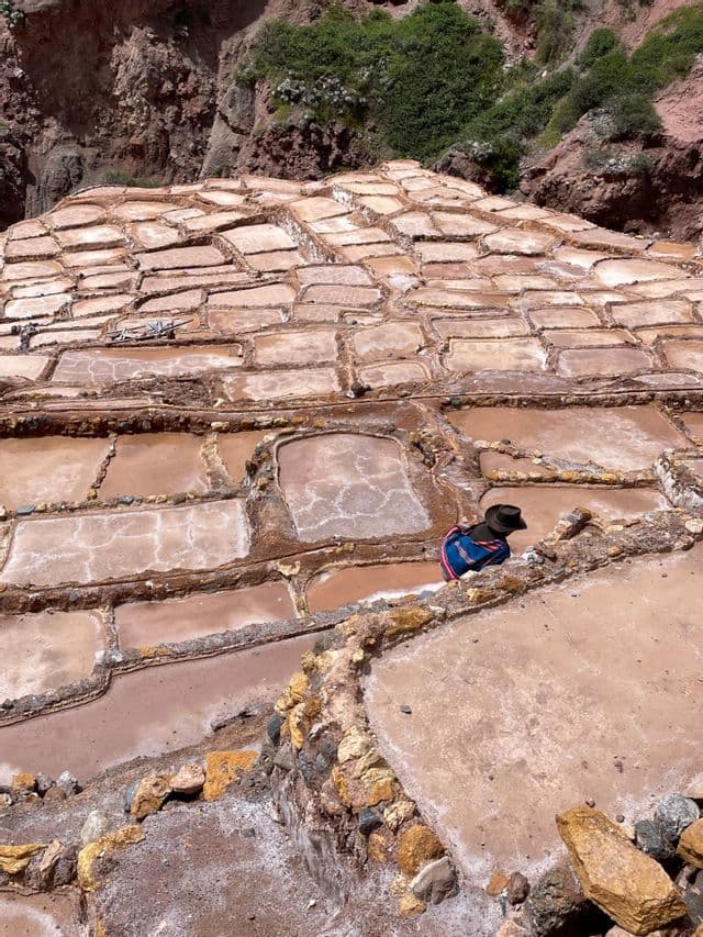 Una persona con un cappello a tesa larga siede sul bordo delle saline terrazzate su un fianco di montagna.