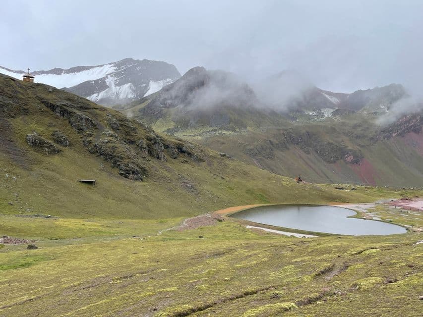 Un piccolo lago in una verde valle di montagna, con cime innevate parzialmente oscurate da nuvole e nebbia.