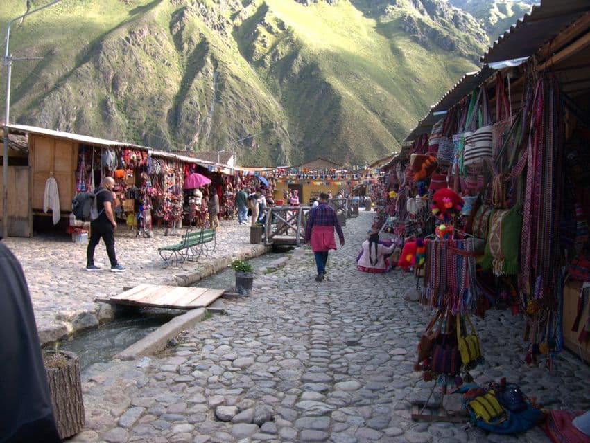 Un mercato all'aperto con bancarelle di tessuti colorati costeggia una strada di ciottoli ai piedi di una grande montagna verde.