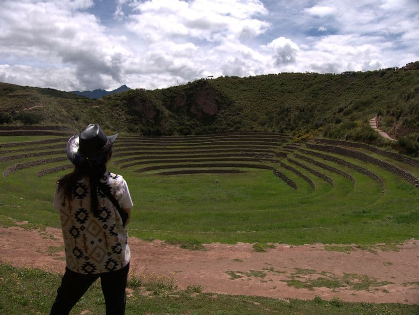 Eine Person mit Hut und gemusterter Weste blickt auf große, kreisförmige landwirtschaftliche Terrassen an einem grünen Berghang.
