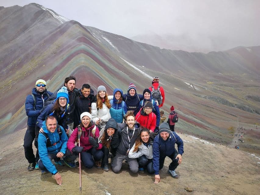 Un gruppo WeRoad in viaggio posa per una foto su una vetta di montagna, con montagne colorate e striate sullo sfondo sotto un cielo nuvoloso.