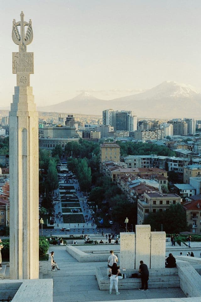 Un monumento in pietra ornato su una terrazza si affaccia su un paesaggio urbano con un ampio viale e montagne innevate in lontananza.