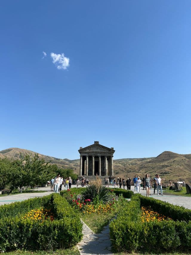 Un antico tempio in pietra con colonne, visibile da un giardino formale, con turisti che passeggiano e colline sullo sfondo sotto un cielo azzurro.