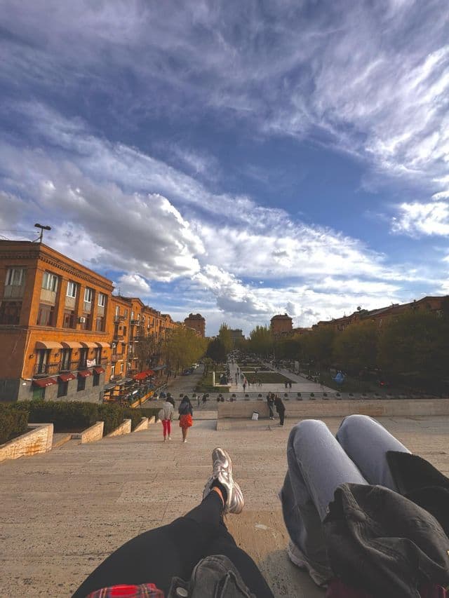 Una vista in prima persona dalla cima di ampie scale di pietra, che guarda giù su una piazza cittadina con edifici arancioni e persone che camminano.