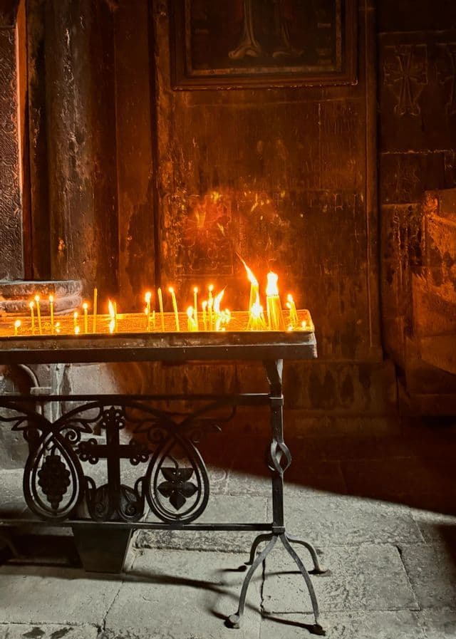 Una fila di candele accese arde su un candelabro in ferro battuto all'interno di una chiesa di pietra scarsamente illuminata.