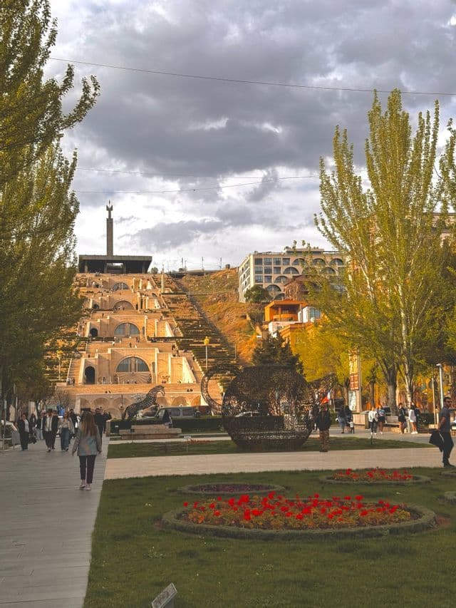 Una grande scalinata in pietra a gradoni sale su un pendio, con un parco, sculture e pedoni in primo piano sotto un cielo nuvoloso.