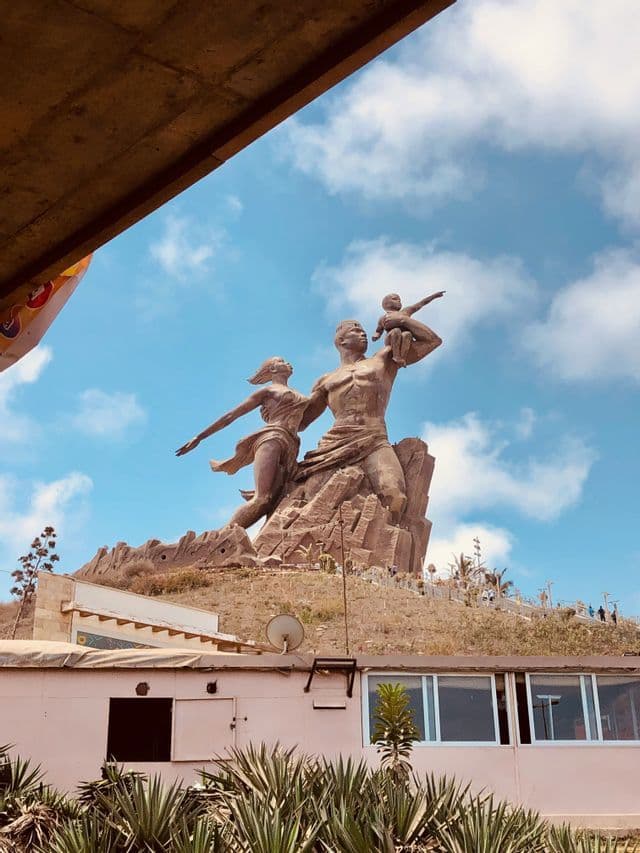 Une statue monumentale en bronze d'une famille se dresse sur une colline rocheuse, sous un ciel bleu avec des nuages, vue en contre-plongée.