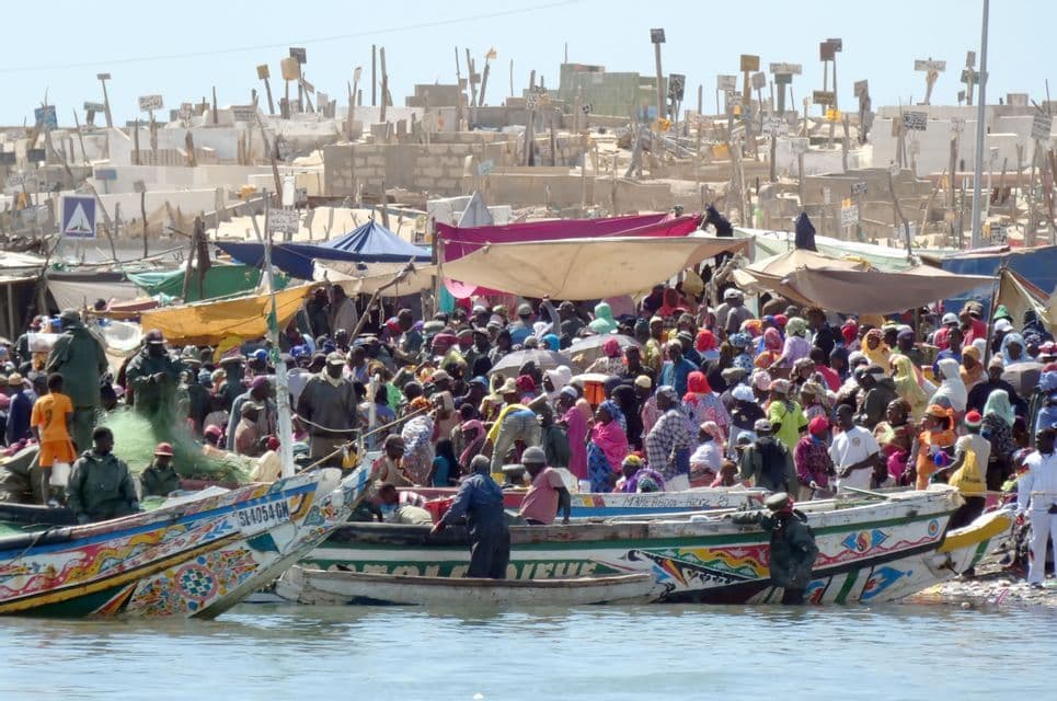 Un front de mer bondé où de nombreuses personnes sont rassemblées autour de bateaux colorés, avec un grand cimetière sur la colline en arrière-plan.