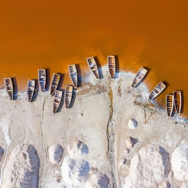 Vue aérienne de bateaux en bois vides reposant sur un rivage de sable blanc au bord d'une étendue d'eau orange.