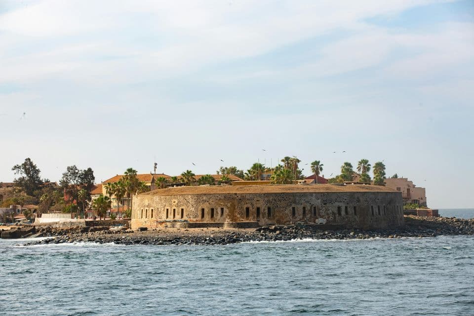 Une forteresse circulaire en pierre se dresse sur un littoral rocheux, vue de la mer, avec des palmiers et des bâtiments en arrière-plan.