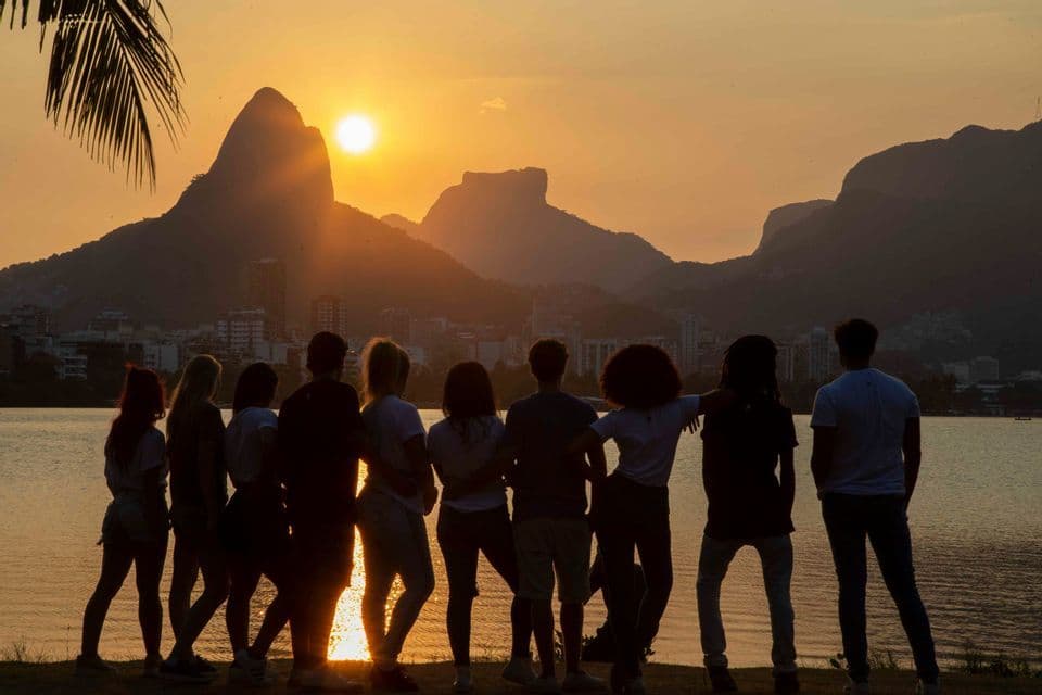 A silhouetted WeRoad group trip stands by a lake, watching the sun set behind mountains and a city skyline.