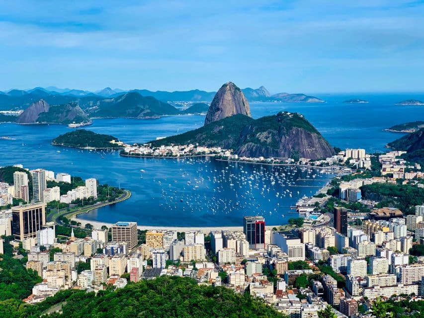 An aerial view of a dense coastal city surrounding a bay filled with sailboats, with large green mountains in the background.