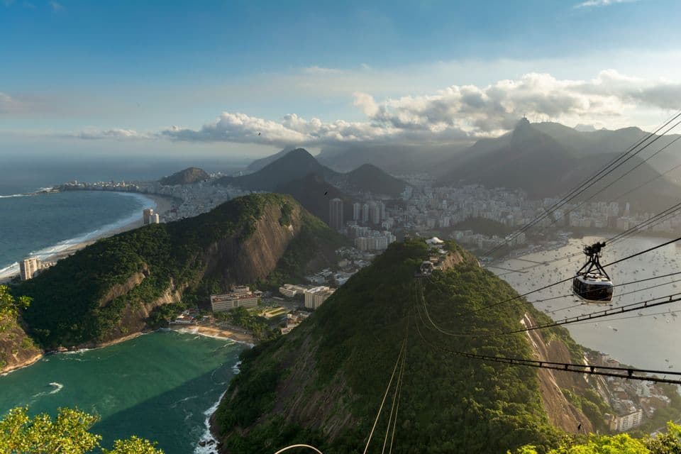 An aerial view of a cable car traveling over lush green mountains, with a coastal city and a bay in the background.