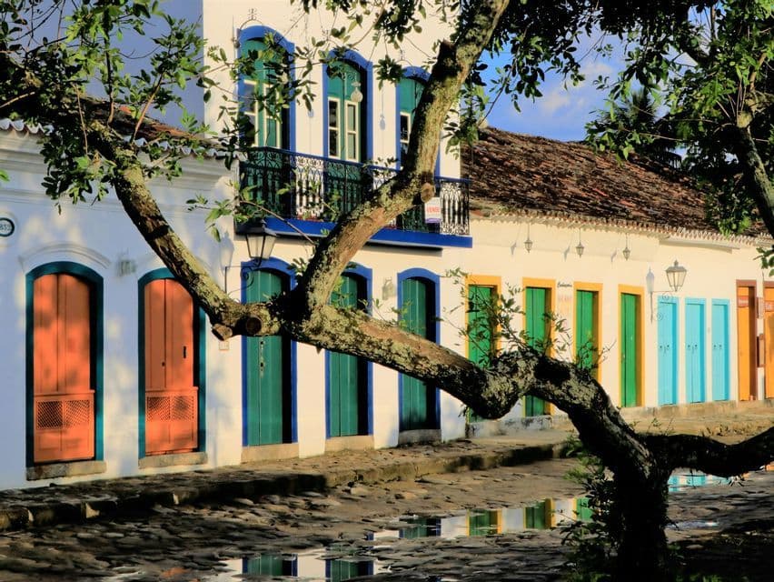 White colonial-style buildings with colorful doors and windows line a wet cobblestone street with a tree in the foreground.