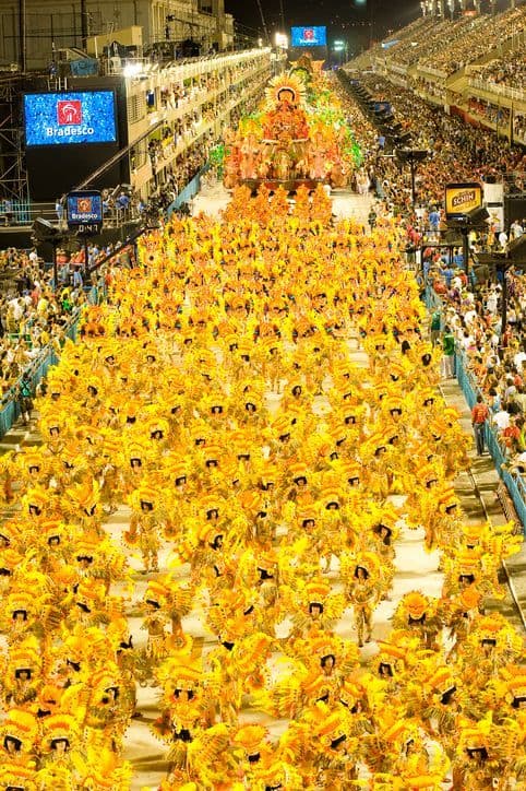 A high-angle view of a nighttime parade with hundreds of performers in bright yellow costumes, watched by crowds in large stands.