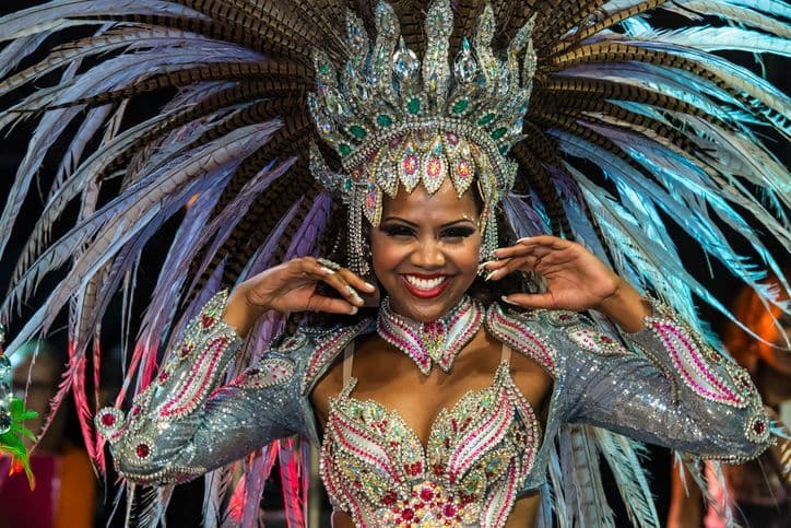 A woman smiles at the camera wearing an elaborate carnival costume with a large, feathered headdress and sparkling jewels.