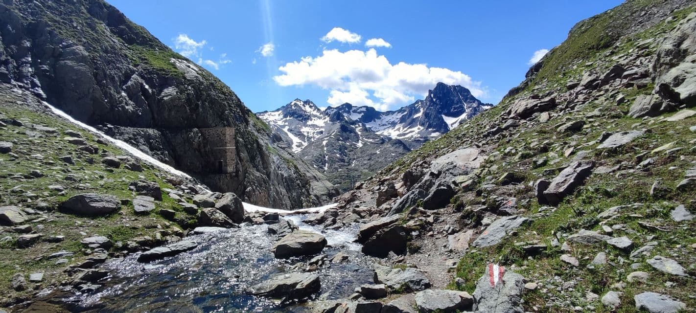Un torrente di montagna roccioso si snoda attraverso una valle con cime innevate in lontananza sotto un cielo azzurro brillante.