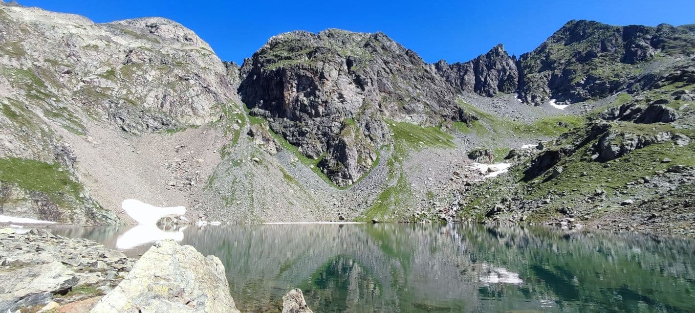 Un limpido lago alpino riflette montagne rocciose e un cielo blu, con chiazze di neve sulla riva.