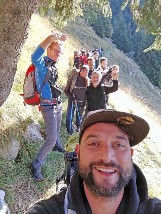 Un uomo si scatta un selfie sorridente con un gruppo WeRoad durante un'escursione in fila indiana su un sentiero di montagna soleggiato.
