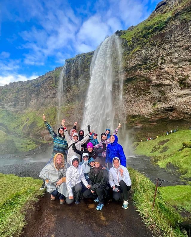 Un viaje en grupo de WeRoad con impermeables sonríe y posa para una foto frente a una gran cascada en un acantilado cubierto de musgo.