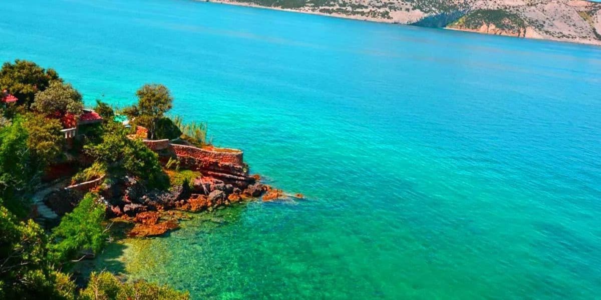 Una veduta dall'alto di una costa rocciosa e verde che si getta in una vasta distesa di mare turchese e blu profondo, con una riva distante sullo sfondo.