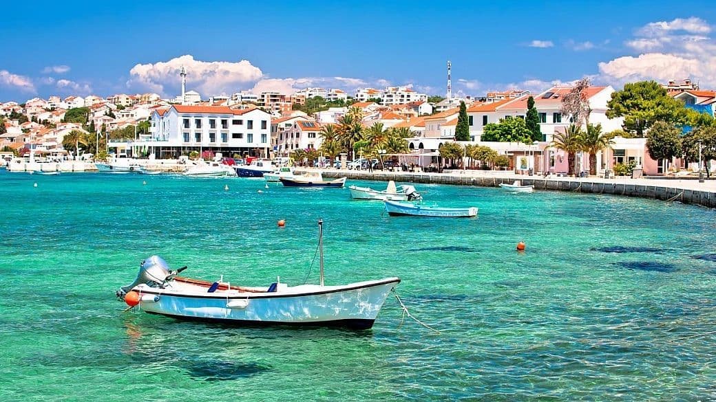 Piccole barche ormeggiate in un porto calmo e turchese con una città costiera illuminata dal sole sullo sfondo sotto un cielo azzurro.