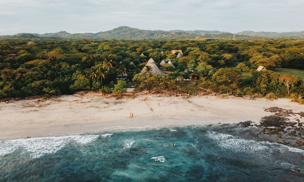 Vista aérea de una playa de arena y océano turquesa junto a un exuberante bosque verde con cabañas de techo de paja y montañas distantes.